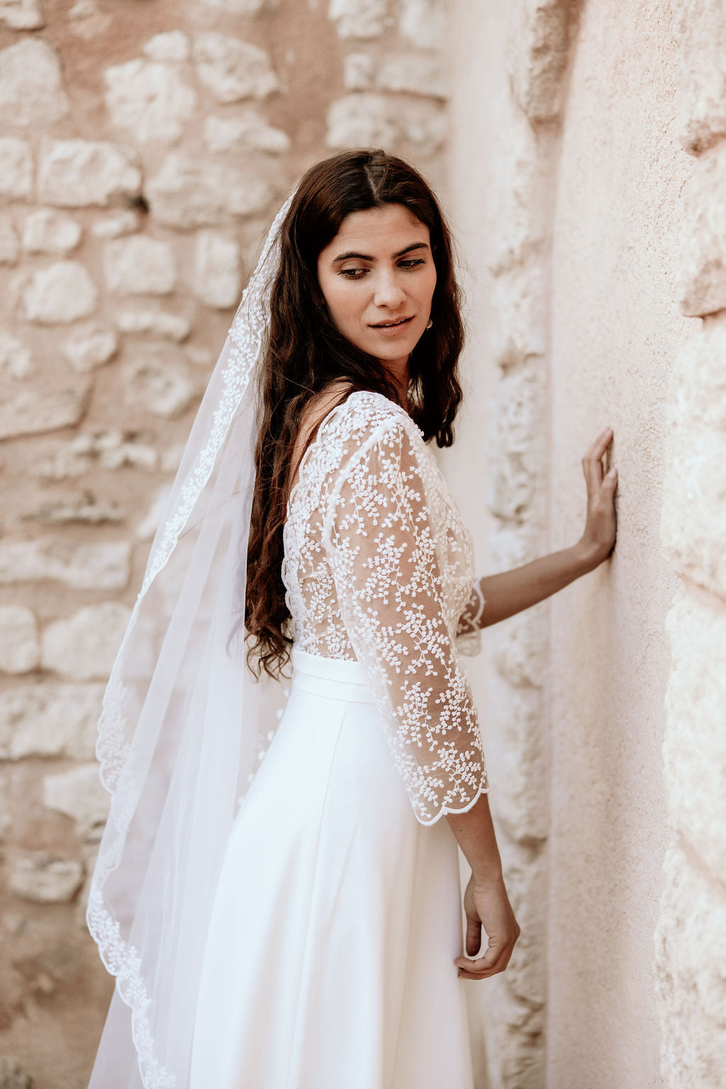 Woman in a white lace dress standing against a stone wall.