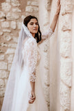 Woman in a white lace wedding dress standing against a stone wall.