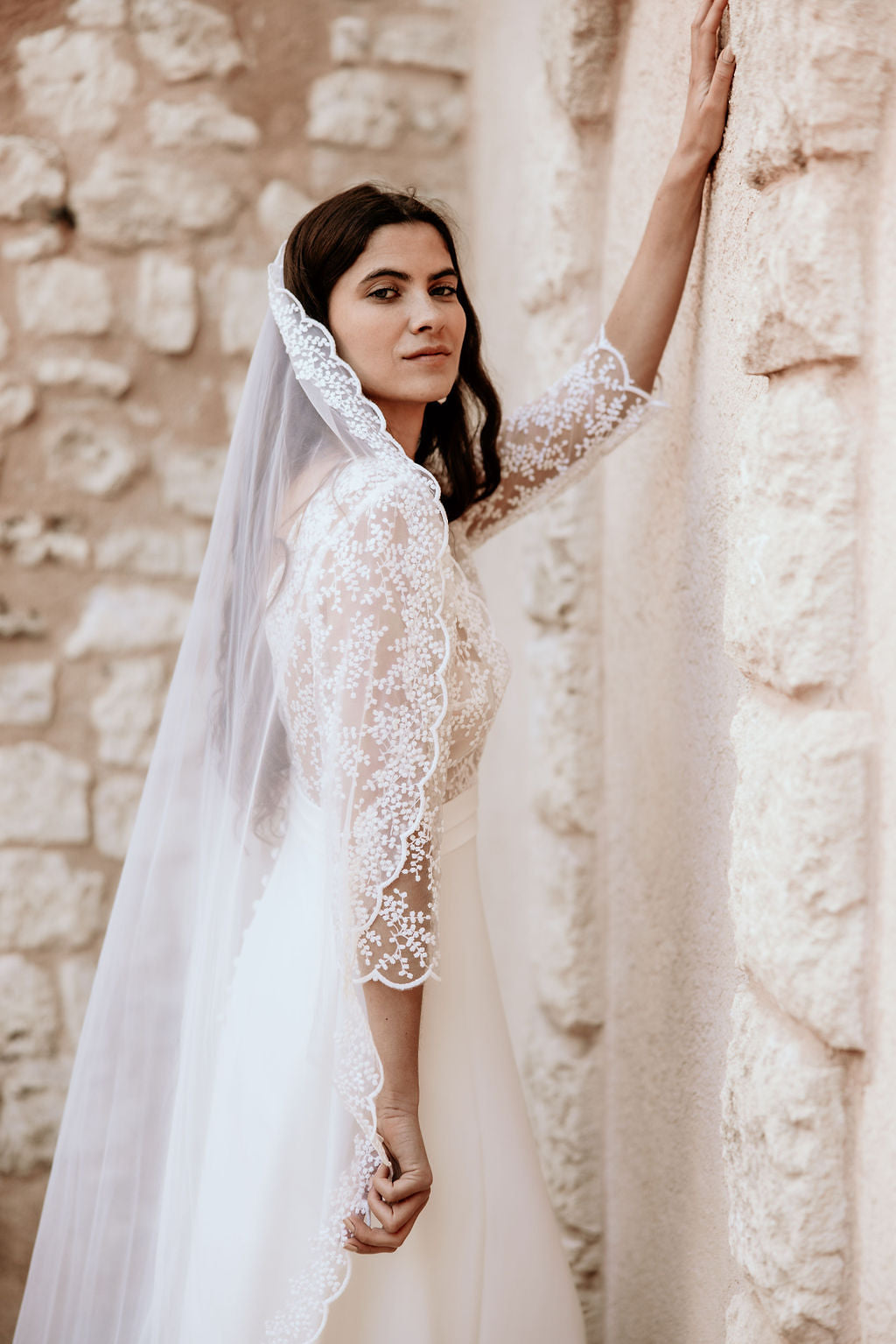 Woman in a white lace wedding dress standing against a stone wall.