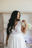 A woman with long dark hair wears a white lace wedding dress, looking to the side. She stands in a bright room with white flowers and a shelf behind her.