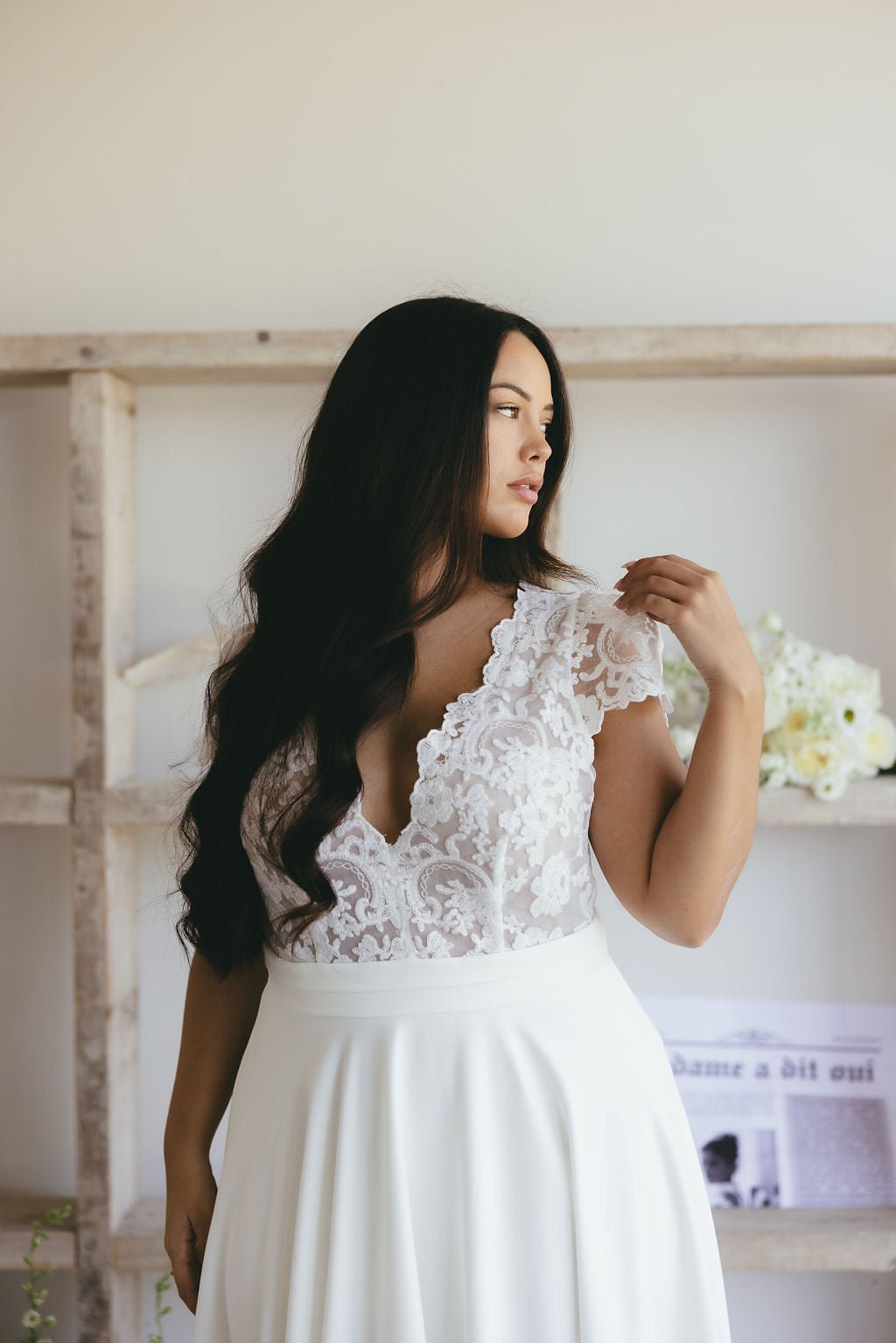 A woman with long dark hair wears a white lace wedding dress, looking to the side. She stands in a bright room with white flowers and a shelf behind her.