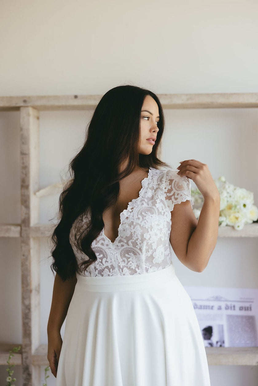 A woman with long dark hair wears a white lace wedding dress, looking to the side. She stands in a bright room with white flowers and a shelf behind her.