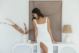 Woman in a white dress sits on a white bench, smiling downwards. Behind her is abstract wall art. A vase with dried leaves and a lamp are nearby.